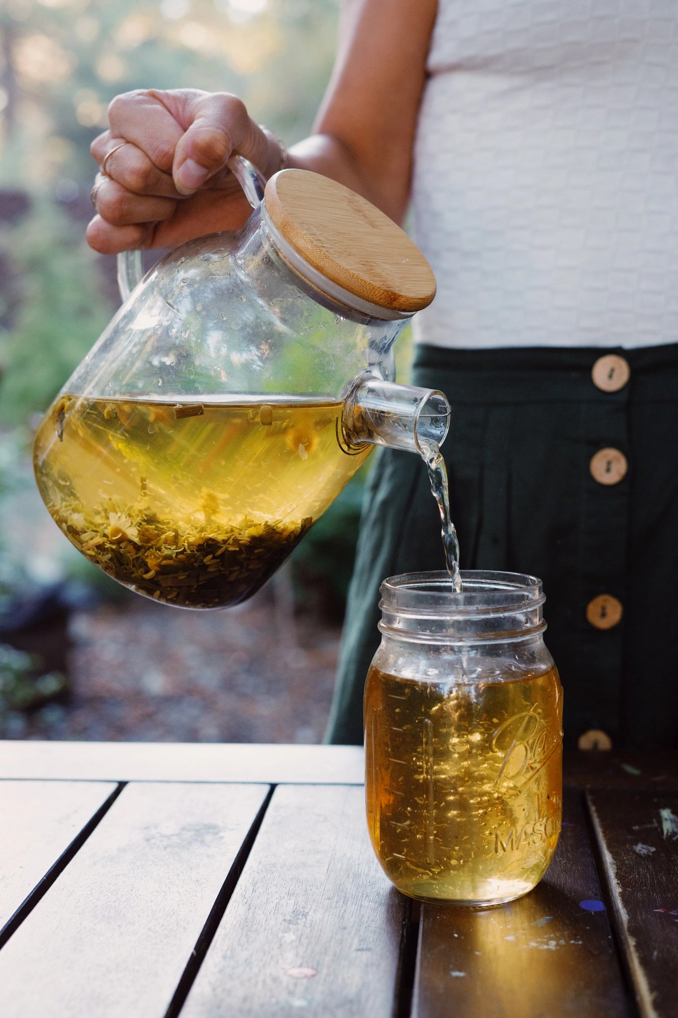 Person pouring tea from a glass teapot with a wooden lid into a mason jar on a wooden table.