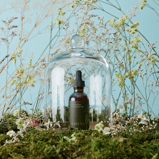 Bottle of SUBLUNA serum under a glass dome with flowers and plants on a light blue background