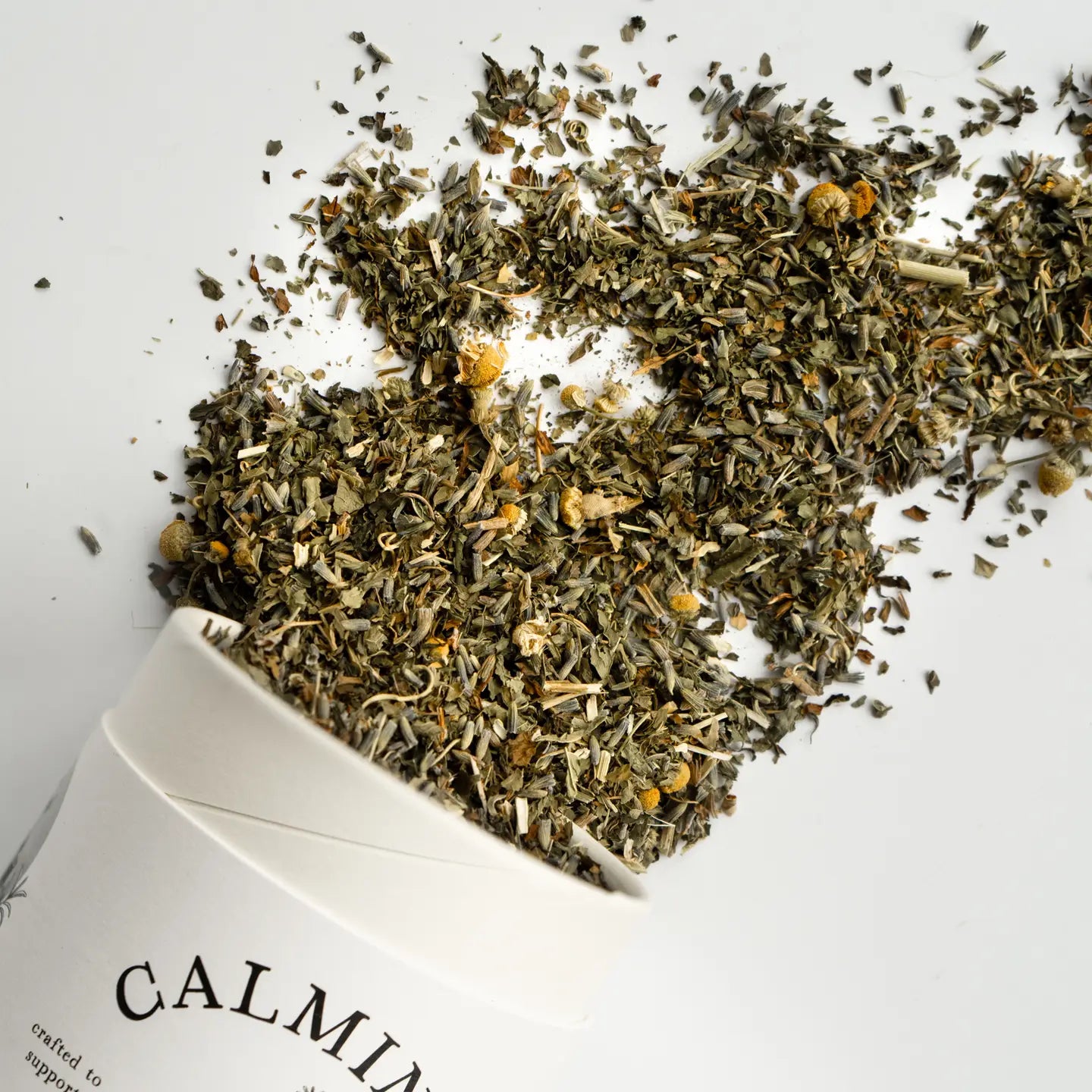 Dried herbs spilling out of a white container labeled 'Calming' on a white background